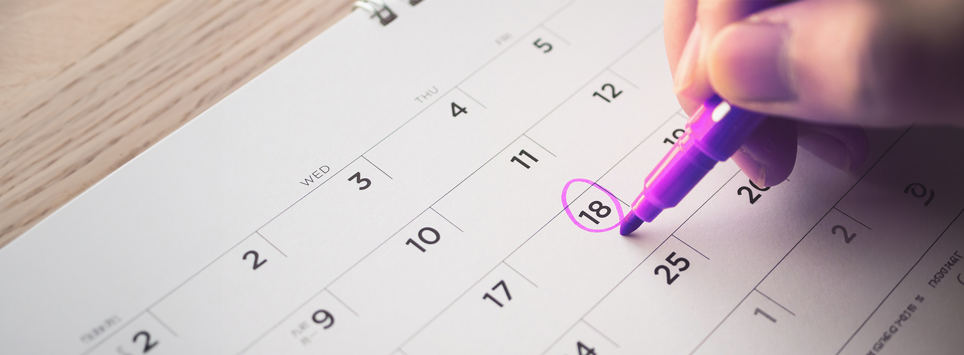 A close-up photograph of a purple pen marking a date on a calendar with a blurred hand holding the pen, set against a wooden surface.