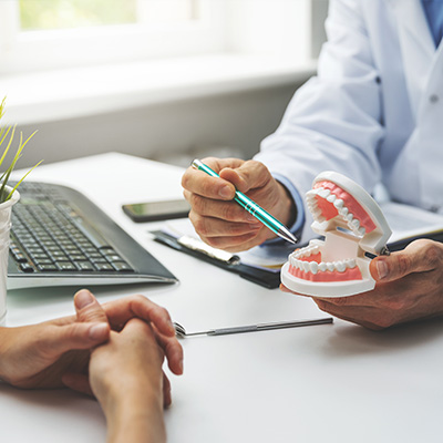 A dental professional examining a patient s teeth model with a penlight, while seated at a desk with a laptop and a potted plant.