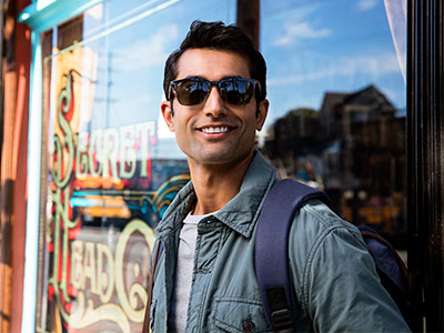 The image shows a man standing outdoors in front of a shop window wearing sunglasses, a backpack, and holding onto his jacket with one hand. He is smiling and looking directly at the camera.