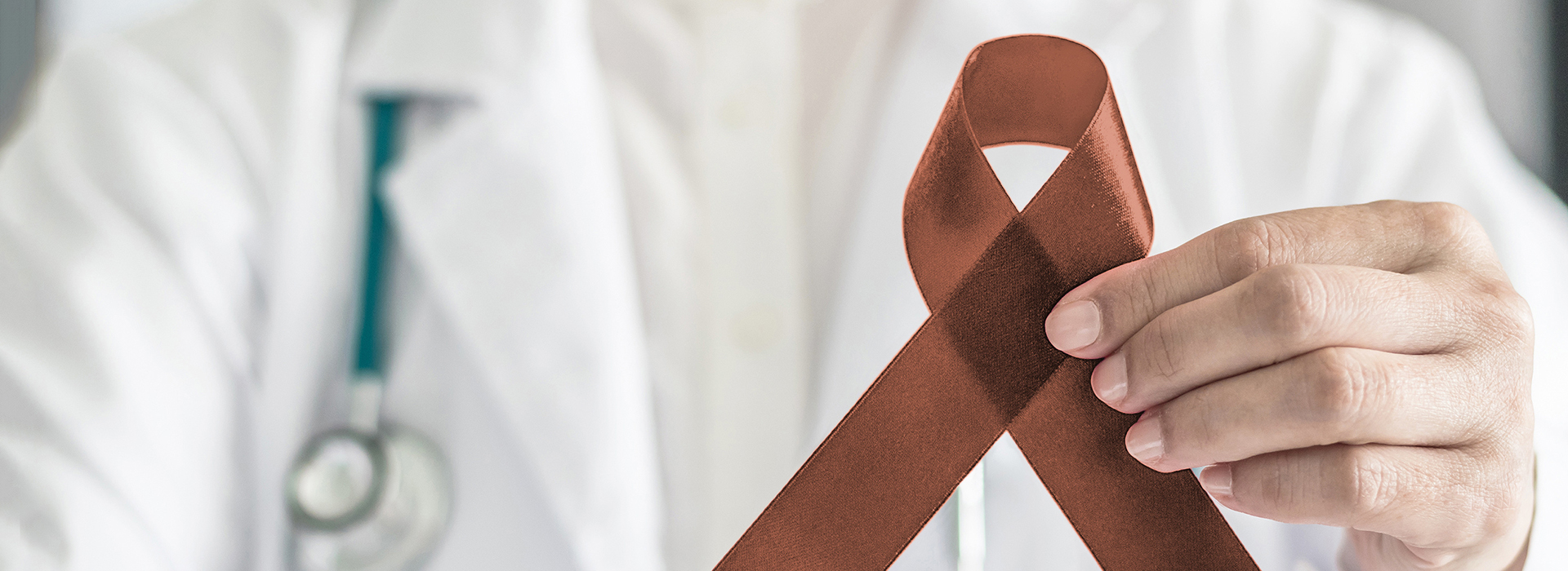 The image shows a person wearing a white lab coat holding up a red ribbon with a brown tie attached to it, symbolizing awareness for a cause  the background is blurred but appears to be an indoor setting.