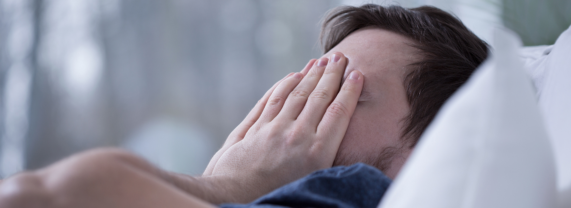 A man lying down on a bed with his face covered by his hands, appearing distressed or upset.