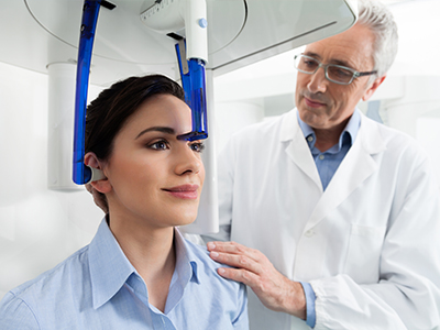 The image shows a woman seated in a dental chair with a device on her head, being attended to by a dentist who appears to be examining or preparing for a procedure.