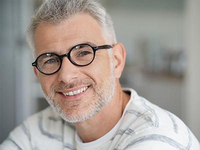 The image shows a man with gray hair, wearing glasses, smiling at the camera. He has a beard and mustache and is dressed casually with a patterned shirt.