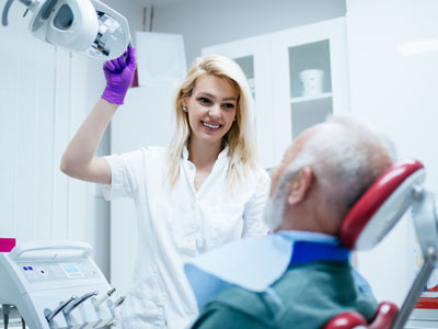 A female dental hygienist assisting an elderly man with dental care equipment in a dental office setting.