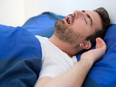 The image shows a man lying in bed with his eyes closed, appearing to be asleep or possibly unconscious, with a blue blanket covering him partially.