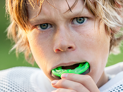 An image of a young boy with blonde hair, wearing a football jersey, holding an object with green and blue colors near his mouth.